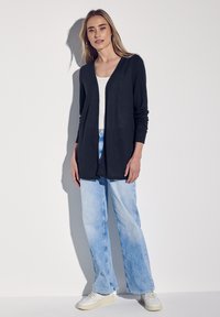 Svart, öppen cardigan med långa ärmar, kombinerad med ljusblå vida jeans och vita sneakers. Slät textur, avslappnad design.