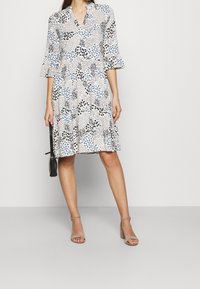 Kek dress berpolakan bunga berwarna krim dengan aksen biru dan hitam. Memiliki leher V, lengan bell pendek, dan hem mengembang. Dipadukan dengan sepatu hak tinggi bertali.