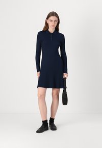 Marineblå ribbestrikket kjole med lange ermer og krage, med en tettsittende bodice og lett utforming nederst. Kombinert med svarte sko og veske.