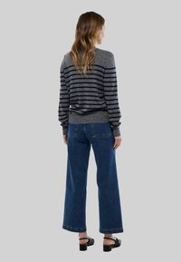 Femme aux cheveux longs portant un pull rayé gris et noir, un jean large bleu et des sandales à talons noires, tournée de dos sur un fond gris.