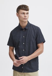 Camisa de botones de manga corta, azul oscuro, en una tela texturizada, con un cuello clásico y botones beige. Combinada con pantalones beige.