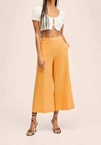 Kobieta w białym krótkim topie i pomarańczowych, wysokich szerokich spodniach, nosząca czarne sandały na obcasie, stoi na tle jednolitego koloru.