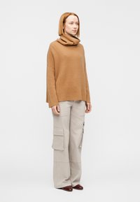 Μπεζ ριγωτό ζιβάγκο πουλόβερ με άνετη εφαρμογή, συνδυασμένο με ανοιχτό γκρι παντελόνια cargo με πολλαπλές τσέπες, και καφέ παπούτσια.