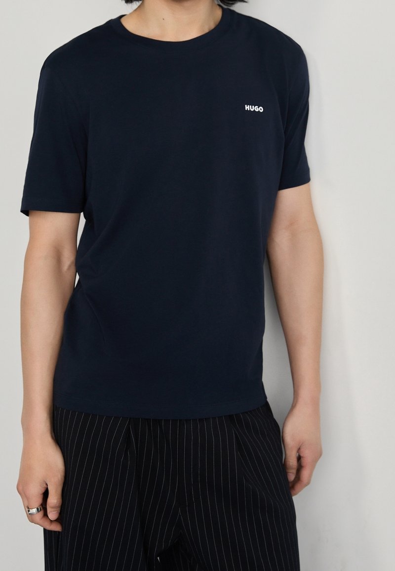 Personne portant un t-shirt Hugo bleu marine et un pantalon noir à fines rayures, debout devant un fond uni gris clair.
