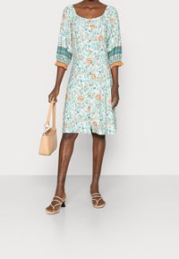 Blommig A-linjekjol i ljusblått med orange och gröna accenter, 3/4 ärmar och fyrkantig halsringning, tillsammans med beige sandaler och en handväska.