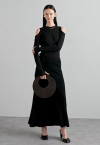 Schwarzes Langarmoberteil mit Ausschnitten an den Schultern, kombiniert mit einem gerippten schwarzen Maxirock. Das Model hält eine runde schwarze Handtasche mit Struktur.