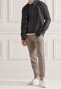 Chemise à boutons gris foncé portée sur un pull sombre, associée à un pantalon chino beige clair et à des baskets blanches avec des accents verts. Textures lisses partout.