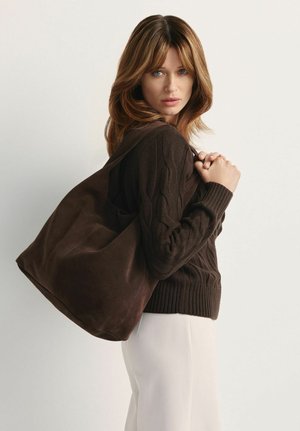 Brązowa zamszowa torba na ramię z szerokimi paskami, miękką teksturą i zaokrąglonym kształtem, pasująca do brązowego swetra z dzianiny i białych spodni.