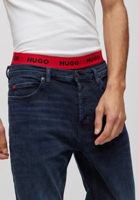 Los jeans azul oscuro cuentan con una cinturilla elástica roja con el logo "HUGO" en negro. Disponibles con bolsillos, textura suave y un diseño ajustado.