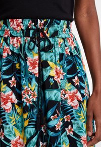 Dettaglio in vita di una gonna con stampa floreale con foglie e fiori tropicali, caratterizzata da un laccio nero e la mano di una persona accanto ad essa.