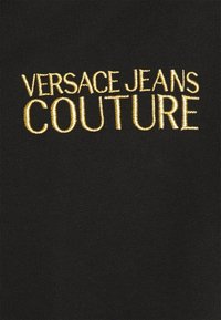 Tela negra con "VERSACE JEANS COUTURE" bordado en hilo dorado. Textura suave con letras claramente definidas.