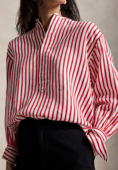 Žena v voľnej červeno-bielej košeli s vertikálnymi pruhmi zapnutej na gombíky, zastrčenej do čiernych nohavíc, otočená mierne do strany.