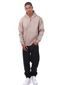 Haut de survêtement zippé beige clair avec une grande capuche et des poches avant, associé à un pantalon noir ample et des baskets blanches, offrant une texture de tissu lisse.