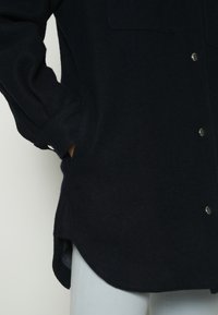 Personne portant un manteau noir boutonné avec la main dans la poche latérale, assorti d'un pantalon de couleur claire, visible de la mi-cuisse au milieu du torse.