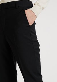 Schwarze, maßgeschneiderte Hosen mit glatter Oberfläche, ausgestattet mit Fronttaschen und schmaler Passform. Der Bund hat Gürtelschlaufen.