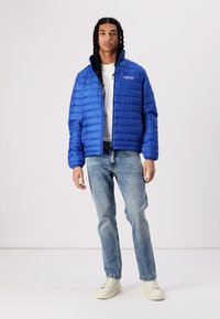 Blauer Pufferjacke mit gestepptem Textur, kombiniert mit hellblauen Jeans und cremefarbenen Sneakers, ausgestattet mit einem Reißverschluss und stehendem Kragen.