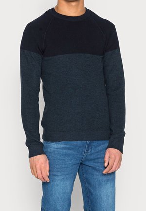 Maglione a costine blu scuro con spalla a contrasto in colore uniforme, scollo tondo e polsini aderenti, abbinato a jeans di denim azzurro chiaro.