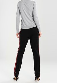 Kvinna klädd i svarta byxor med röda sidostreck, grå långärmad topp och svarta högklackade sandaler, stående med ryggen vänd.