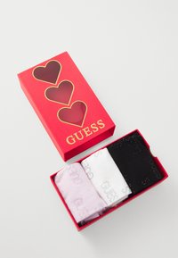 Tri parova čarapa u crvenoj kutiji s izrezanim srcima. Čarape su ružičaste, bijele i crne s otisnutim "GUESS" u metalik boji.