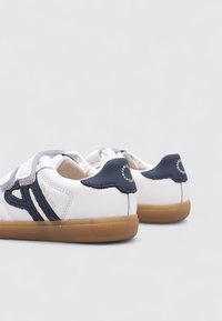 Zapatillas blancas con acentos en azul marino, con un upper de cuero suave, correas de Velcro y una suela de goma. Marca notable en el talón.