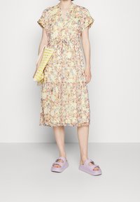Femme portant une robe midi à fleurs pastel, tenant un sac tissé jaune et blanc, avec des sandales à plateforme lilas, sur un fond uni.