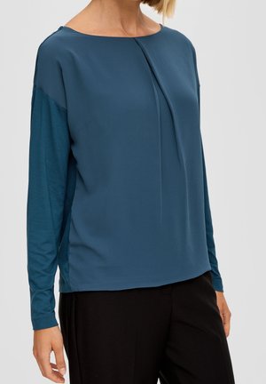 Kobieta ubrana w turkusową, długorękawową bluzkę z plisowanym przodem, w połączeniu z czarnymi spodniami na jasnoszarym tle.