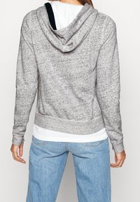 Person iført en meleret grå hættetrøje over en hvid skjorte og lyseblå jeans, vist fra ryggen mod en ensfarvet baggrund.