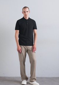 Mann som står mot en grå vegg, iført en svart polo-skjorte, beige cargo-bukser og hvite sneakers, og ser rett frem.