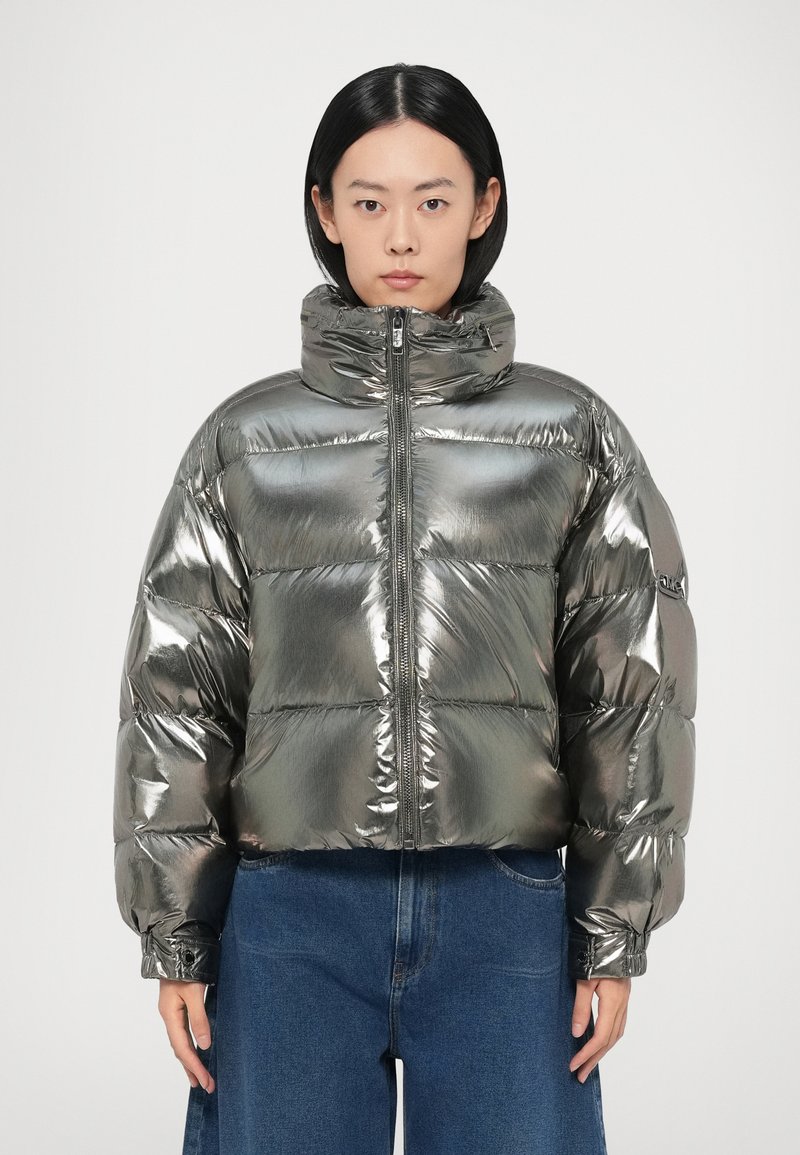 Metāliskas sudraba krāsas pūkaina jaka ar augstu apkakli, šūtu dizainu, priekšēju rāvējslēdzēja aizdari un elastīgām aprocēm, kombinēta ar zilu džinsu audumu.