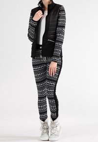 Gilet imbottito nero indossato sopra una maglietta bianca, abbinato a leggins neri con un motivo geometrico bianco. Stivali bianchi con la cima alta completano l'outfit.