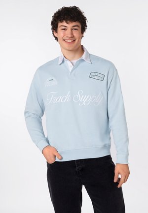 Lächelnder junger Mann mit lockigem Haar, der einen hellblauen Sweatshirt mit dem Schriftzug „Track Supply“ und schwarze Hose trägt, steht vor weißem Hintergrund.