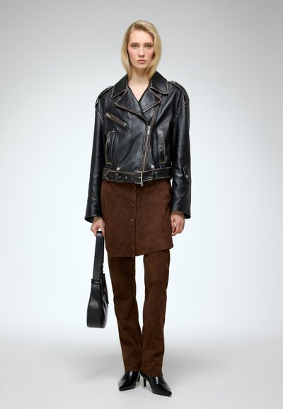 Black leather motorcycle jacket with brown suede skirt and matching pants; holding a black handbag; pointed black heels.