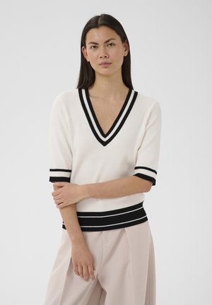 Woman with long dark hair wearing a white V-neck sweater with black stripes and light beige wide-leg pants, posing with arms crossed.