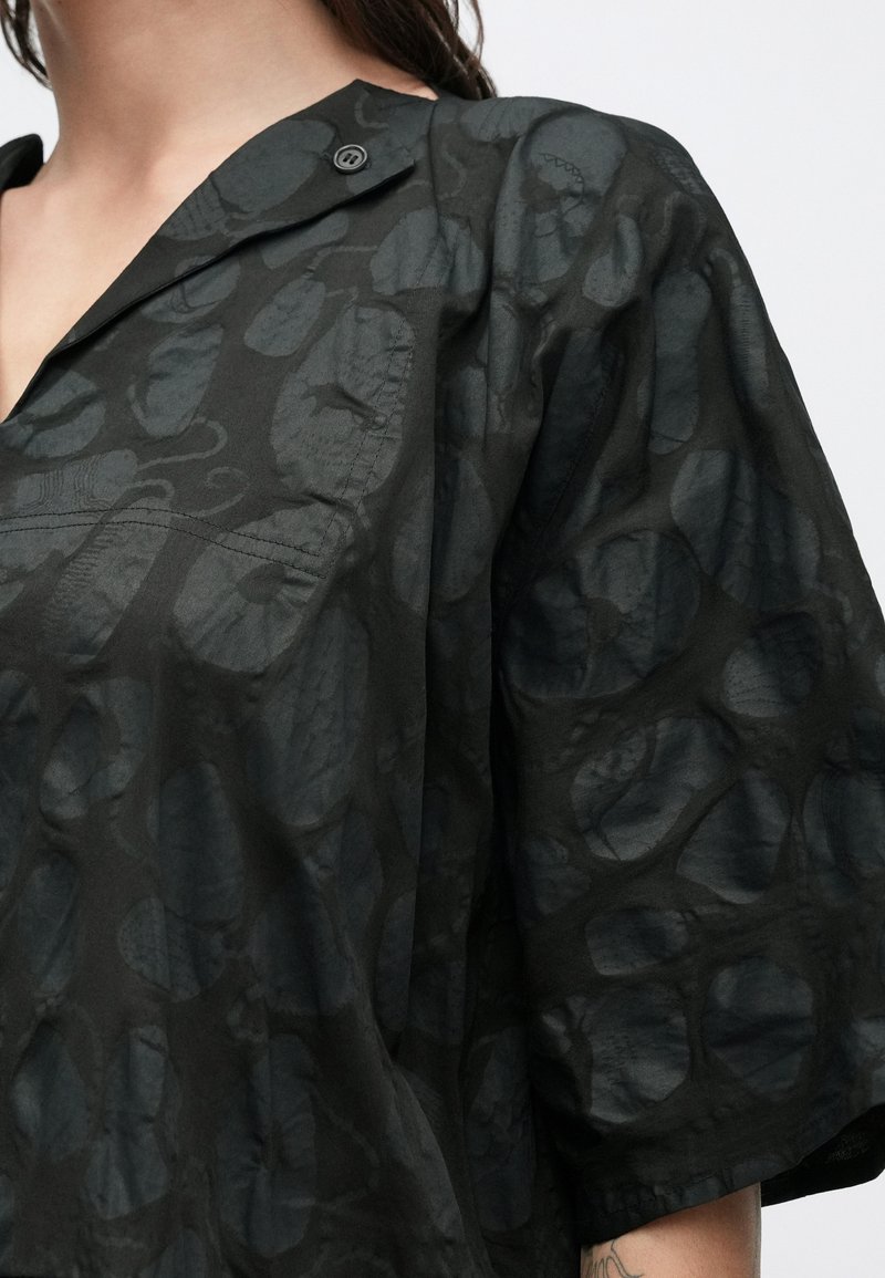 Chemise noire avec un motif texturé. Elle possède un col, des manches courtes et une fermeture par boutons. Le tissu est légèrement transparent et léger.