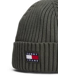 Tumši zaļš adīts cepure ar ribotu tekstūru. Tās priekšpusē ir neliels aplis ar "TOMMY JEANS" un sarkano, baltu un zilo logo.