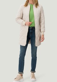 Giacca trapuntata beige con cerniere laterali, polsini e colletto crema, indossata sopra un maglione verde strutturato e jeans blu scuro, abbinata a sneakers beige.