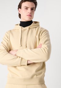 Hanorac bej cu glugă, dotat cu șnururi de ajustare, manșete și tiv cu nervuri. Prezintă un logo subtil pe piept, o textură moale și o croială lejeră.