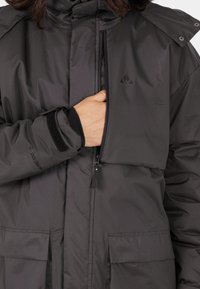 Veste isolante grise avec un col montant, fermeture zippée à l'avant, poches à soufflet et poignets en Velcro. Avec un logo sur la poitrine et un tissu résistant.