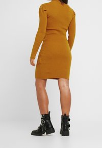 Žluté žebrované pletené šaty s dlouhými rukávy a nabíranými detaily na ramenech, doplněné černými kotníkovými botami s přezkami a masivní podrážkou.