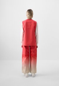 Rote Gradient-Weste und weite Hose, mit einem subtilen Ombre-Effekt von Rot zu Beige, aus weichem Stoff gefertigt. Weiße Sneakers runden den Look ab.