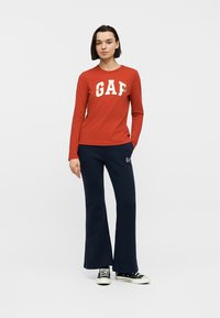 Femme debout, la main dans la poche d'un pantalon bleu marine, portant un pull rouge à manches longues avec le texte blanc "GAP" et des baskets noires.