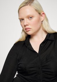 Blusa preta com botões, com colarinho, apresentando um frente pregueada e mangas longas, feita de um tecido texturizado.