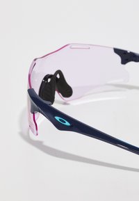 Sportsolglasögon med en marinblå ram, genomskinliga rosa linser och svarta gumminässtöd. Har en blå logodetalj på skalmen.