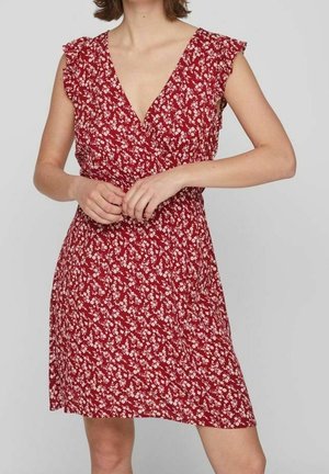 Femme portant une robe rouge à fleurs sans manches avec un col en V et des détails à volants aux épaules, debout devant un fond uni.