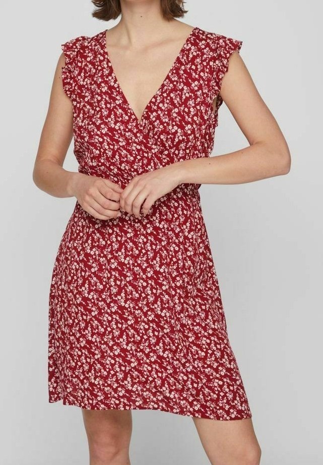 Femme portant une robe rouge à fleurs sans manches avec un col en V et des détails à volants aux épaules, debout devant un fond uni.