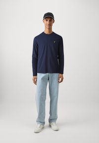 Marineblauw shirt met lange mouwen en een klein geel logo, gecombineerd met lichtblauwe jeans en witte sneakers, aangevuld met een zwarte pet.