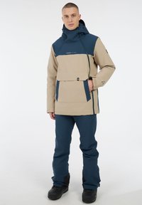 Beigefarbene und marineblaue Softshelljacke mit einer großen Fronttasche, Kapuze und verstellbarem Zugband; kombiniert mit blauen Hosen und schwarzen Stiefeln.