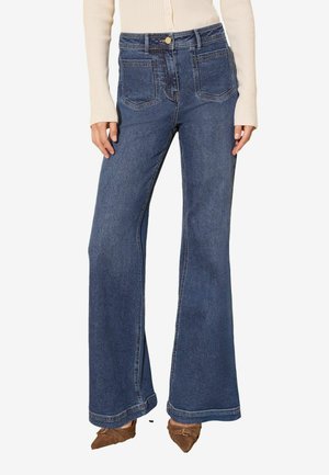 Kvinde iført højtaljede, vidde blå jeans kombineret med brune spidse sko og en beige ribbet langærmet top.