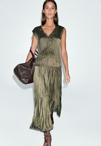 Vestito verde oliva con texture plissettata, scollo a V e maniche corte a mantella, abbinato a una borsa in pelle marrone scuro.