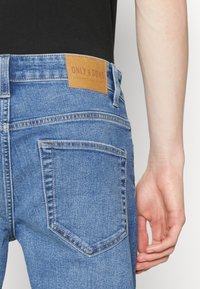 Blå denimjeans med en läderlogga märkt "ONLY & SONS." Har en klassisk femfickorsdesign och synliga sömnadsdetaljer.